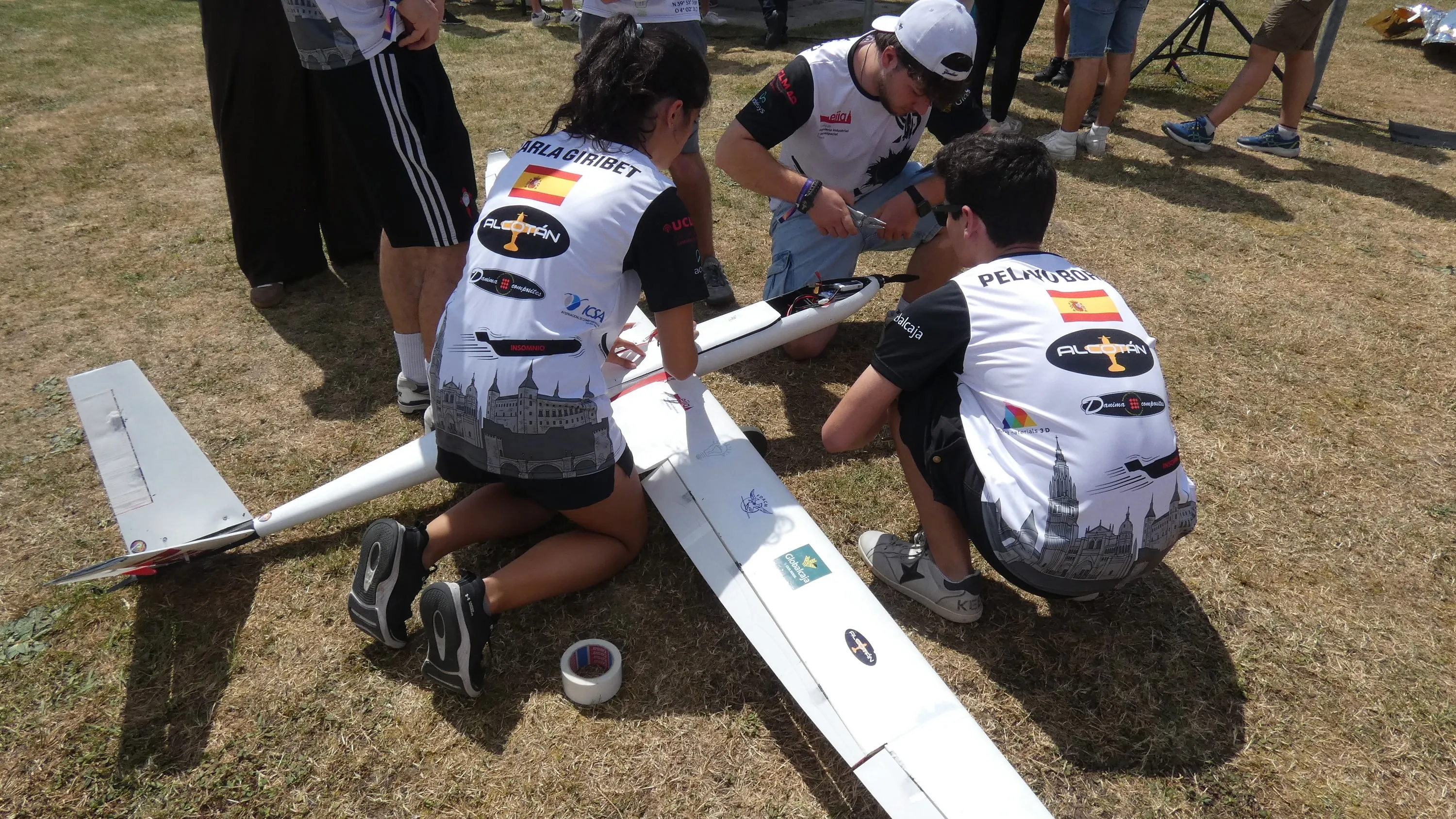 Tres miembros arrodillados en la hierba ajustan el avión, con el dorsal 26 en el ala.