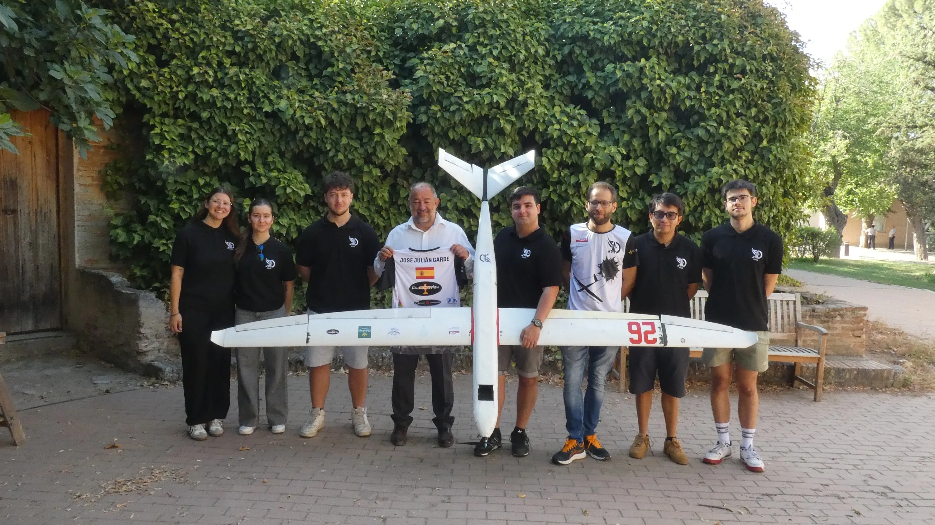 Nueve personas, entre ellas el rector de la UCLM y el profesor tutor del equipo, con el avión blanco del Air Cargo Challenge en un patio con hiedra.