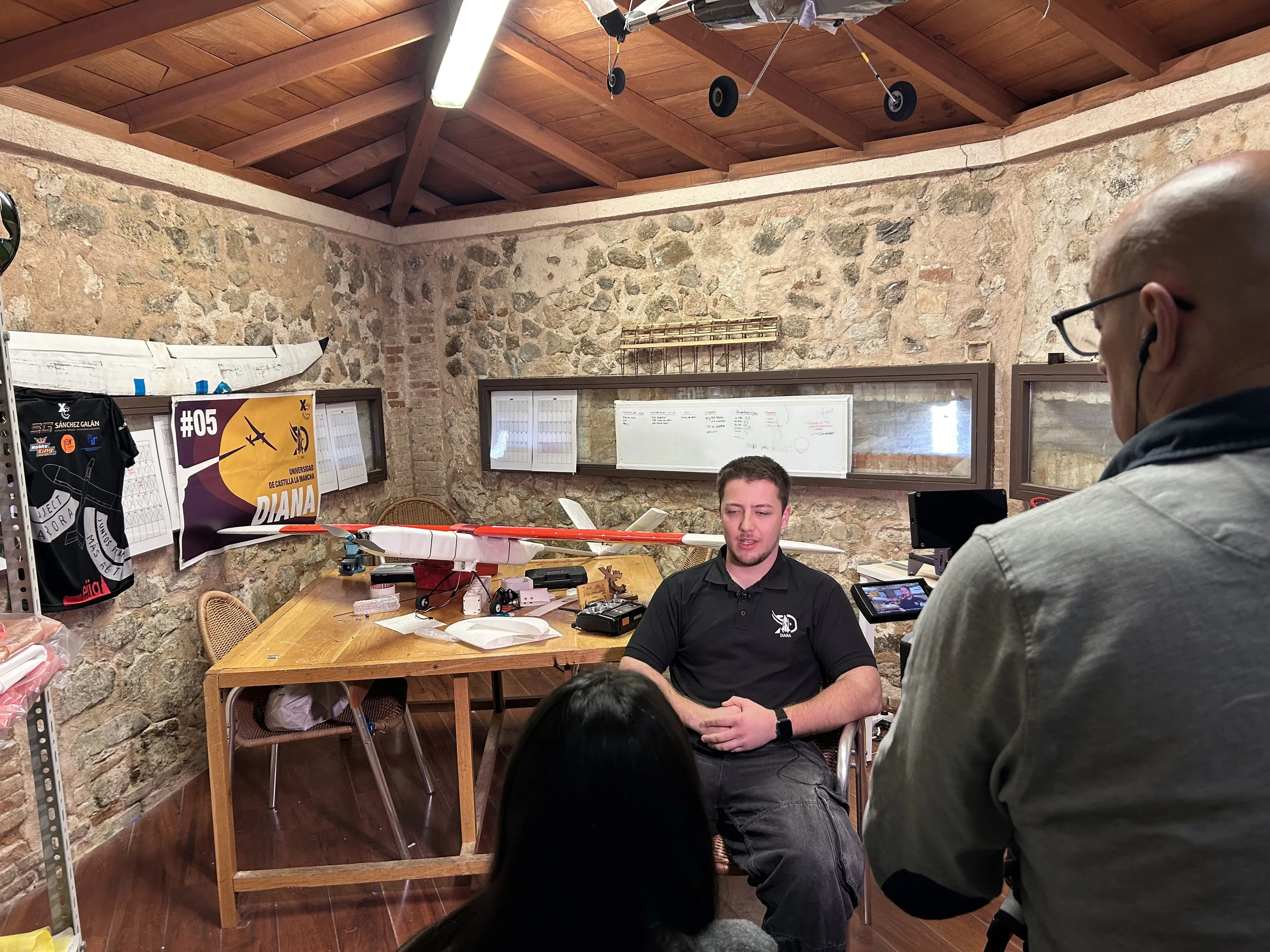 Un miembro del equipo durante una entrevista de televisión, con aeromodelos colgados del techo y una cámara.